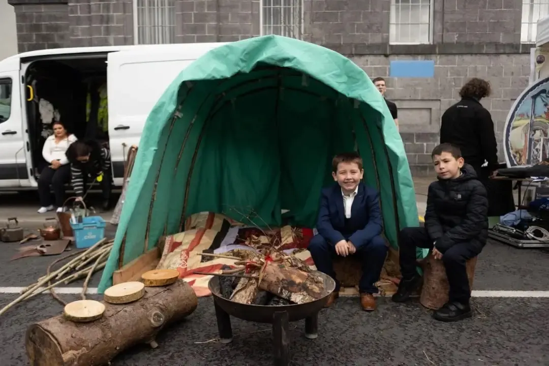 A green canvas shelter, cooking fire and people at an outdoor cultural display.